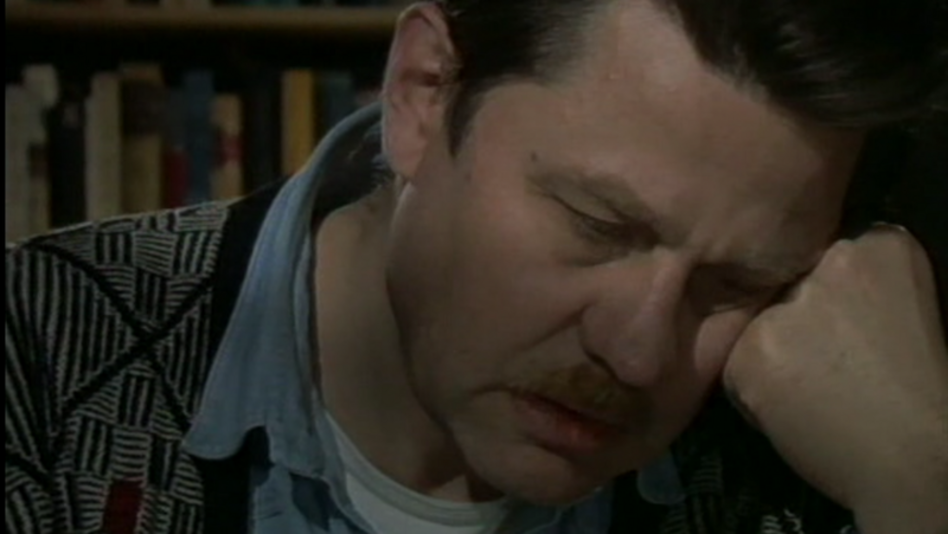 Filmstill aus DAS KINO UND DER TOD: Ein Mann (Hartmut Bitomsky) sitzt an einem Tisch und stützt den Kopf in die Hände. Im Hintergrund ist ein Bücherregal zu sehen.