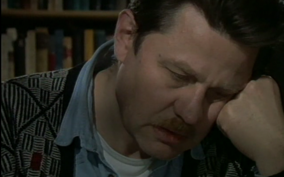 Film still from DAS KINO UND DER TOD: A man (Hartmut Bitomsky) is sitting at a table with his head in his hands. A bookshelf can be seen in the background.