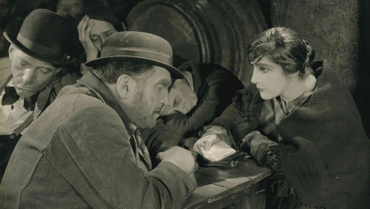 Filmstill aus SCHATTEN DER WELTSTADT: In einer ärmlichen Kneipe beugen sich ein Mann und eine Frau an einem Tisch zueinander. Sie hält ein Bündel Geldscheine in der Hand. Im Hintergrund schlafen einige Männer.