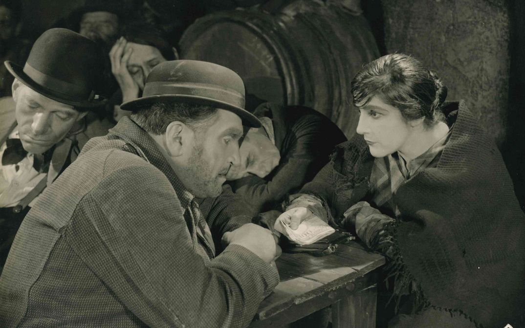 Film still from SCHATTEN DER WELTSTADT: In a pub, a man and a woman bend toward each other at a table. She holds a bundle of banknotes in her hand. In the background, some men are sleeping.