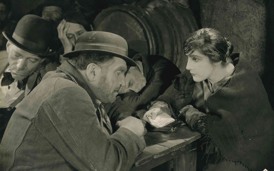 Film still from SCHATTEN DER WELTSTADT: In a pub, a man and a woman bend toward each other at a table. She holds a bundle of banknotes in her hand. In the background, some men are sleeping.
