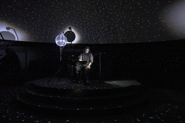 Liz Rosenfeld is sitting on a stage in the planetarium in front of a microphone, the room is dark. Two mirror balls are illuminated and reflect many small points of light in the room