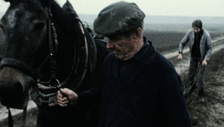 Filmstill aus dem Film "Ein Herbst im Ländchen Bärwalde" von Gautam Bora. Ein Mann mit dunkler Jacke und Mütze führt auf einem matschigen Feld ein Pferd neben sich. Hinter ihnen schiebt eine Person einen Pflug.