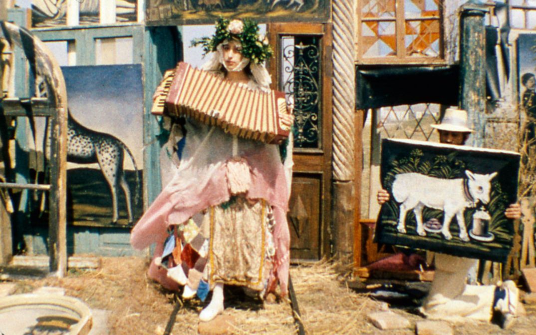 Filmstill aus ARABESKEN ZUM THEMA PIROSMANI: Eine Frau in einem extravaganten Kostüm spielt Akkordeon. Im Hintergrund sind, an Türen und Fenster gelehnt, mehrere Gemälde Pirosmanis zu sehen.