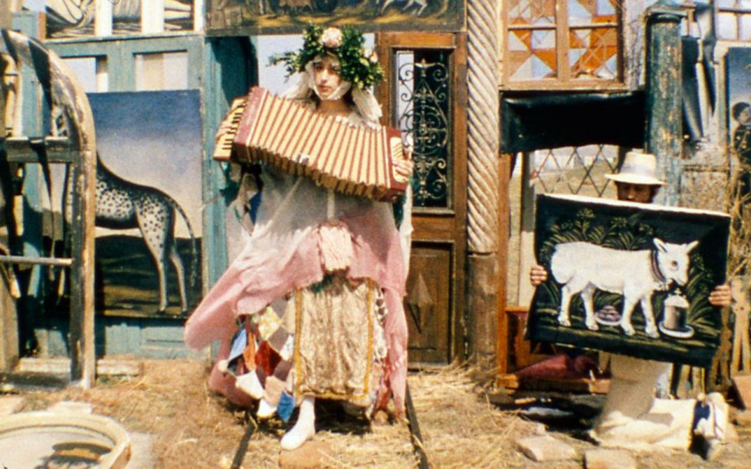 Film still from ARABESKEN ZUM THEMA PIROSMANI: A woman in an extravagant costume plays the accordion. In the background, leaning against doors and windows, several paintings by Pirosmani can be seen.