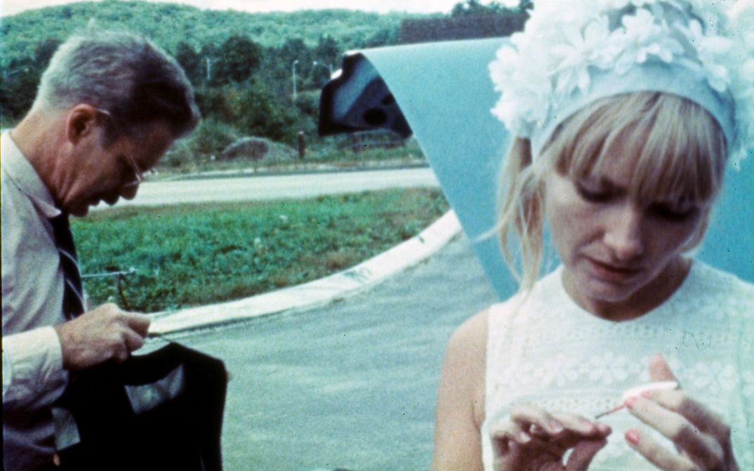 Filmstill aus WANDA: A woman in a flowered bonnet paints her nails. A man stands next to her. There is a street in the background.