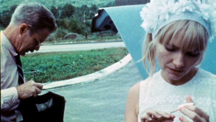Filmstill aus WANDA: A woman in a flowered bonnet paints her nails. A man stands next to her. There is a street in the background.