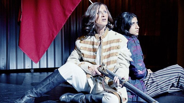 Still from the film "L’état et moi" by Max Linz. Two women in fantasy uniforms sit on a stage. One holds an old-fashioned rifle in her hand. 