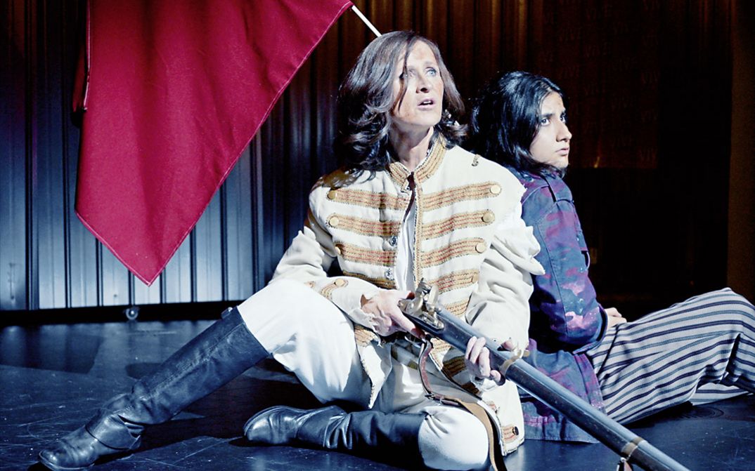 Still from the film "L’état et moi" by Max Linz. Two women in fantasy uniforms sit on a stage. One holds an old-fashioned rifle in her hand. 