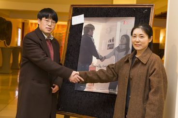 A man and a woman are shaking hands looking into the camera. They are standing in front of a poster, which is a photo of them. In the photo, they are also shaking hands. 