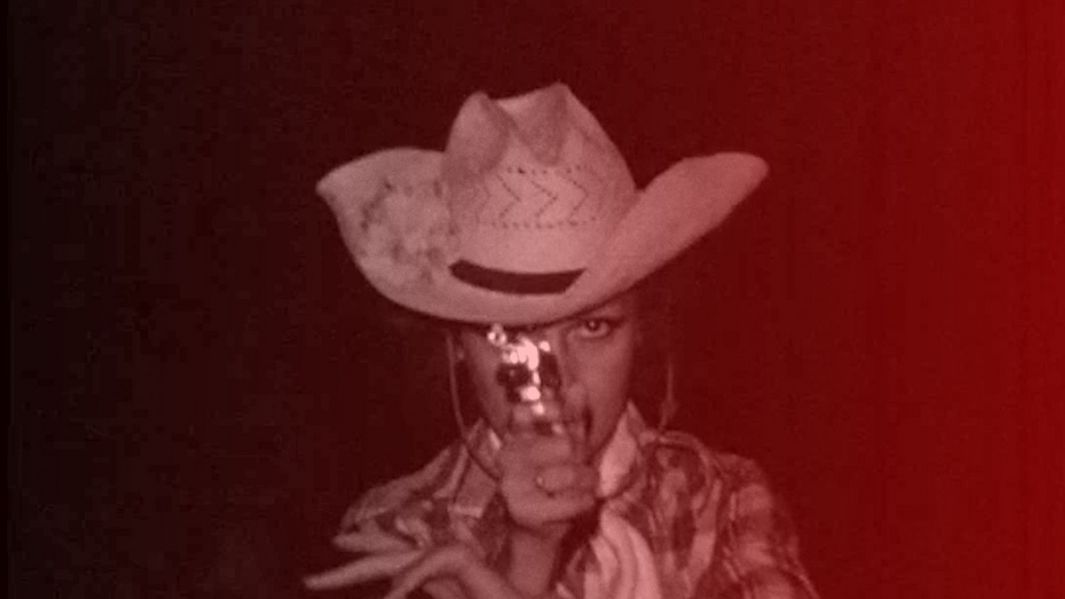 Filmstill aus BAJO LAS BANDERAS, EL SOL: Eine Person in Cowboyhut und Hemd richtet in theatralischer Art einen Revolver auf die Kamera. Das Bild ist monochrom dunkel-rötlich gefärbt.