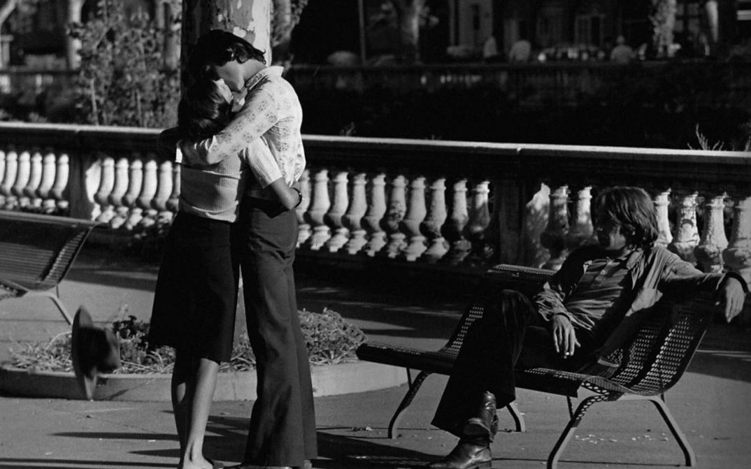 Film still from MES PETITES AMOUREUSES: A girl and a boy are standing close together in a park and kissing. To the right, a boy sits casually on a park bench and watches them.