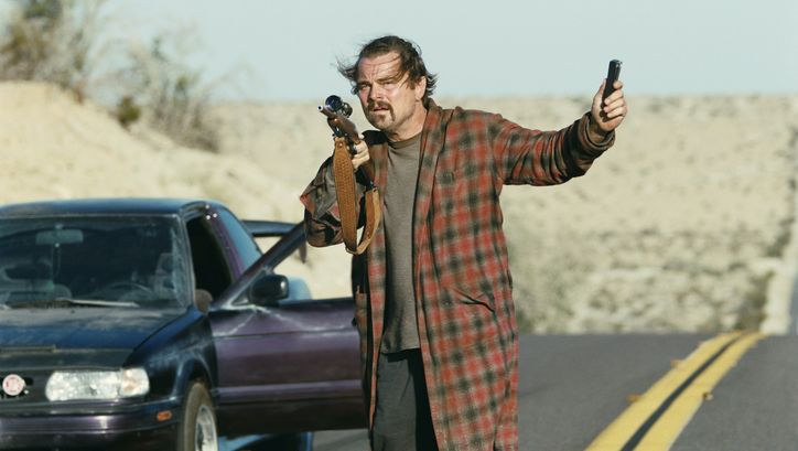 Film still from ONE BATTLE AFTER ANOTHER: A disheveled-looking man in a bathrobe is standing by the road next to the open door of a car. He is holding a rifle in his hand.