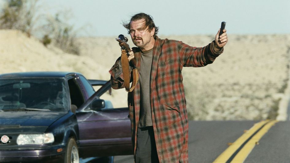 Filmstill aus ONE BATTLE AFTER ANOTHER: Ein zerzaust aussehender Mann in einem Bademantel steht an der Straße neben der offenen Tür eines Autos. In der Hand hält er ein Gewehr.