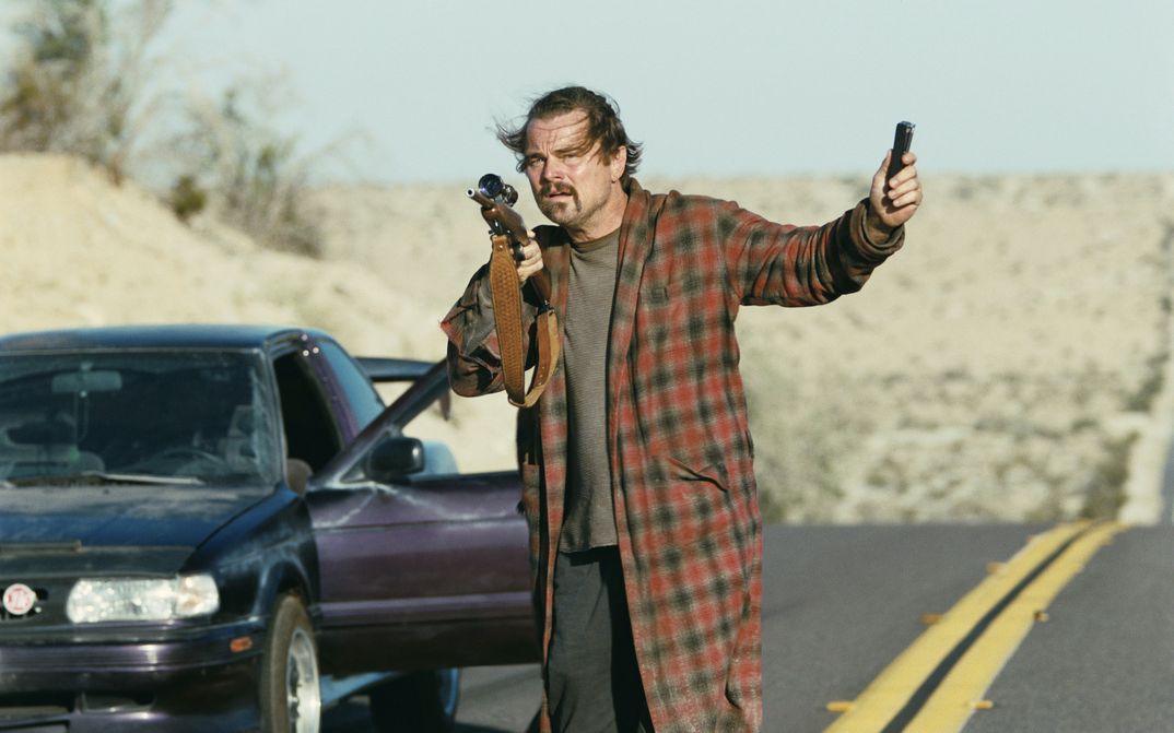 Film still from ONE BATTLE AFTER ANOTHER: A disheveled-looking man in a bathrobe is standing by the road next to the open door of a car. He is holding a rifle in his hand.