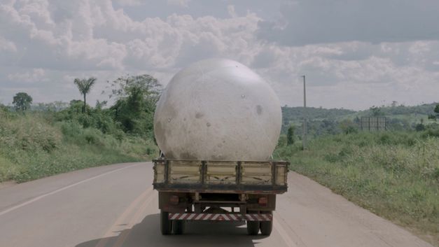 Film still from Janaina Wagner’s film “QUEBRANTE”. A large moon balloon is transported on a pickup truck on a sunny road. 