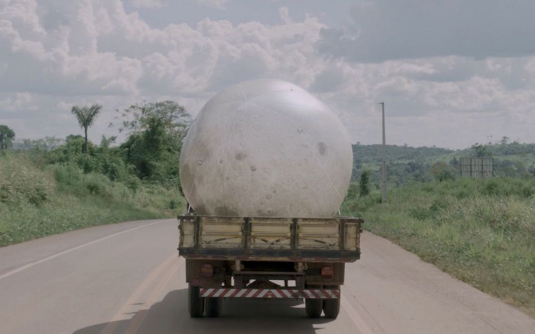 Film still from Janaina Wagner’s film “QUEBRANTE”. A large moon balloon is transported on a pickup truck on a sunny road.