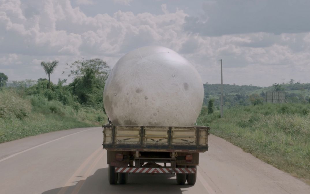 Film still from Janaina Wagner’s film “QUEBRANTE”. A large moon balloon is transported on a pickup truck on a sunny road.
