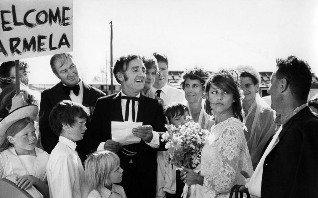 Filmstill aus BELLO, ONESTO, EMIGRATO AUSTRALIA, SPOSEREBBE COMPAESANA ILLIBATA: Eine Gruppe von Menschen umringt ein Brautpaar, jemand hält ein Schild mit der Aufschrift "Welcome Carmela".