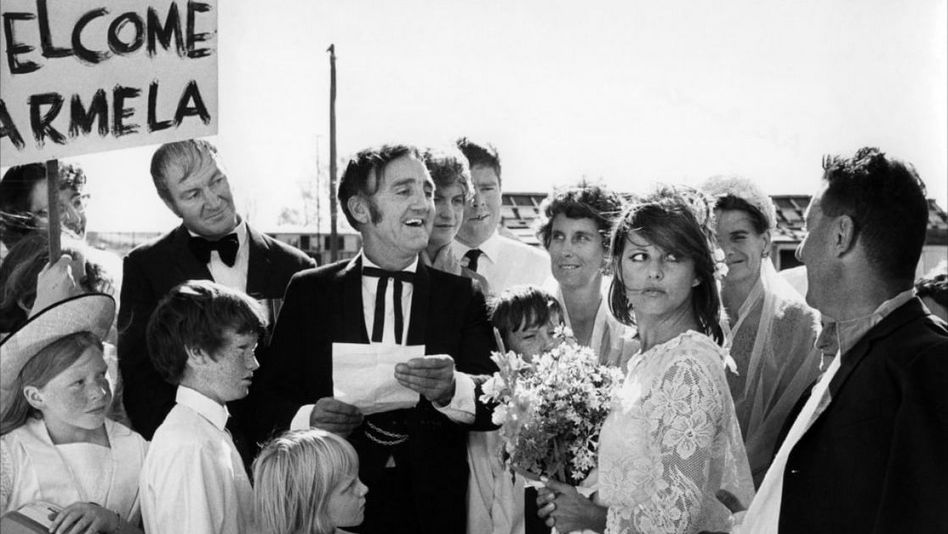 Filmstill aus BELLO, ONESTO, EMIGRATO AUSTRALIA, SPOSEREBBE COMPAESANA ILLIBATA: Eine Gruppe von Menschen umringt ein Brautpaar, jemand hält ein Schild mit der Aufschrift "Welcome Carmela".