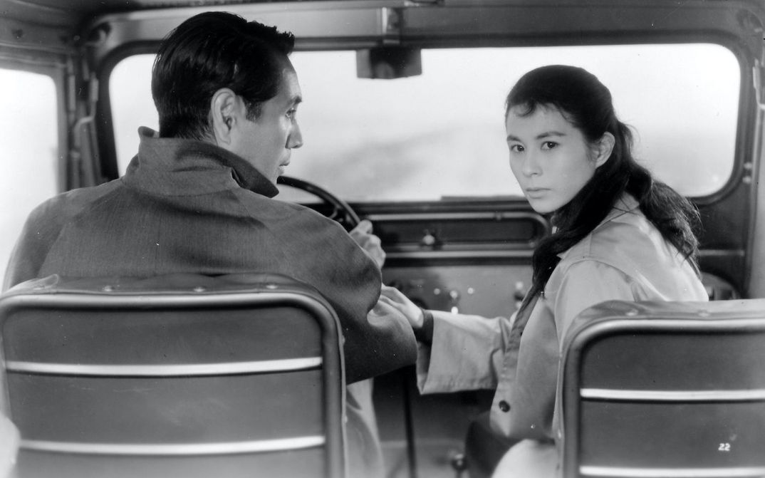Filmstill aus ELEGY OF THE NORTH: Ein Mann und eine Frau sitzen in einem Auto, er ist am Steuer. Sie dreht sich nach hinten um, er sieht sie an.