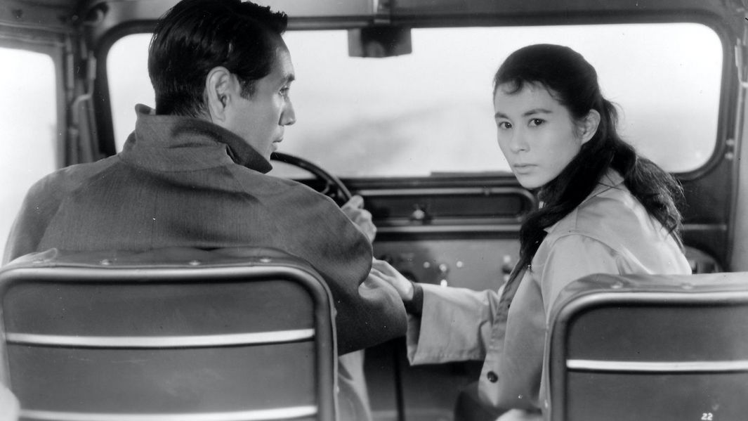 Filmstill aus ELEGY OF THE NORTH: Ein Mann und eine Frau sitzen in einem Auto, er ist am Steuer. Sie dreht sich nach hinten um, er sieht sie an.