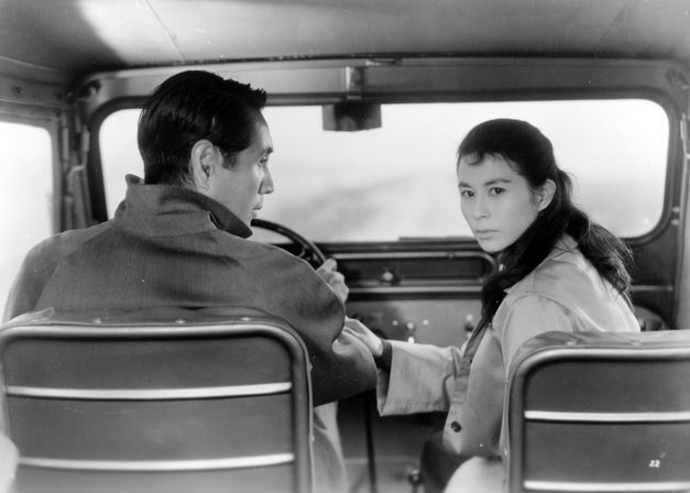 Filmstill aus ELEGY OF THE NORTH: Ein Mann und eine Frau sitzen in einem Auto, er ist am Steuer. Sie dreht sich nach hinten um, er sieht sie an.