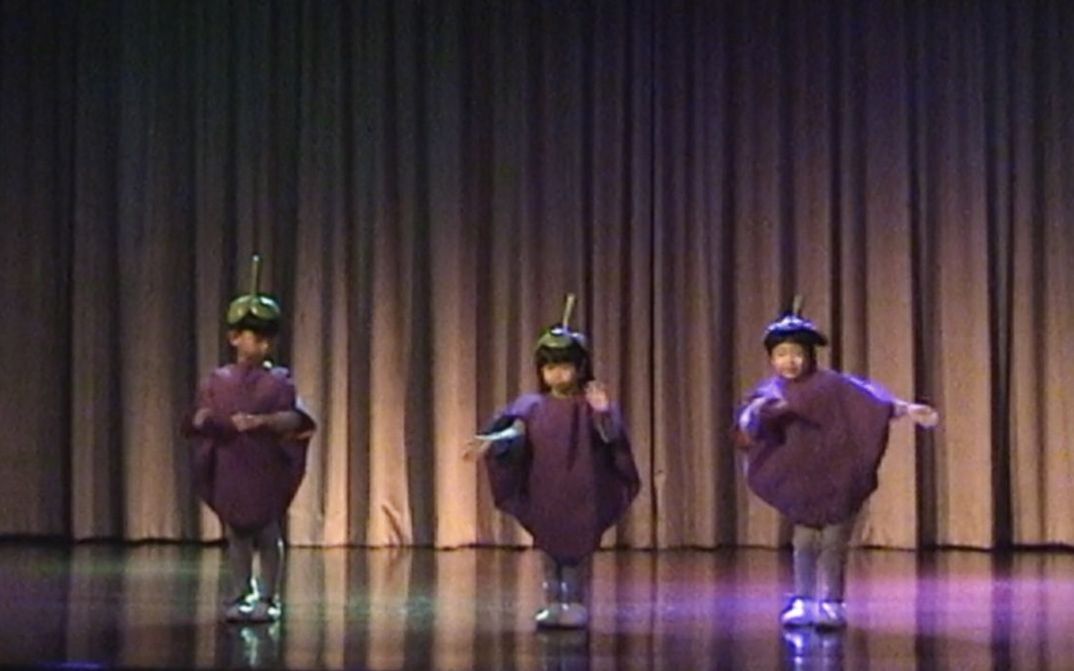 Filmstill aus dem Film „Mangosteen“ von Tulapop Saenjaroen. Drei Kinder in identischen Fruchtkostümen nebeneinander auf einer Bühne. Im Hintergrund ein Vorhang.