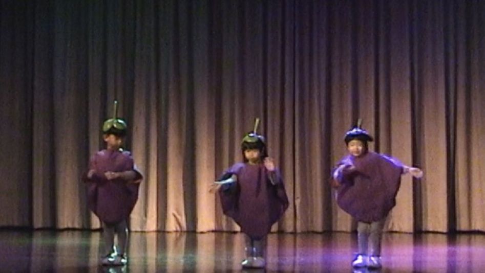 Filmstill from Tulapop Saenjaroen’s „Mangosteen“. Three children in identical fruit-costumes next to each other on a stage. A curtain in the background.