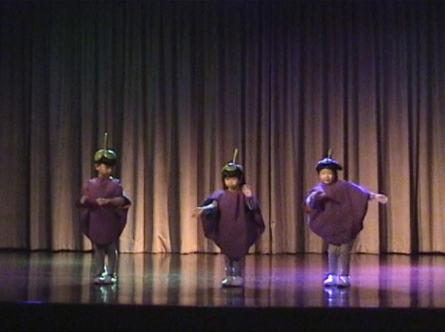 Filmstill from Tulapop Saenjaroen’s „Mangosteen“. Three children in identical fruit-costumes next to each other on a stage. A curtain in the background.