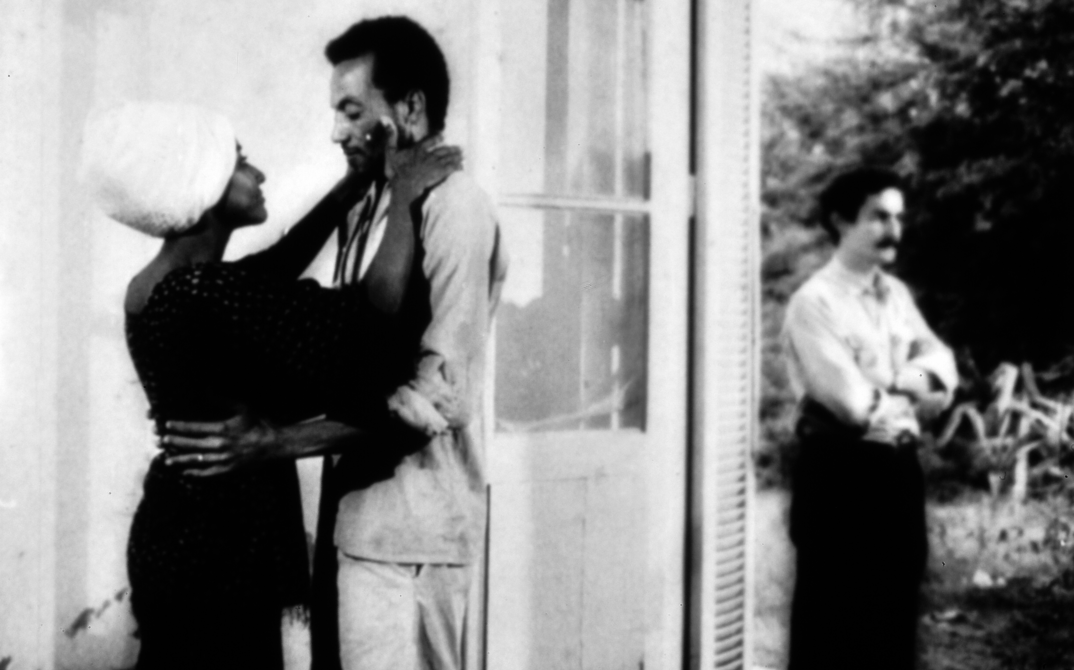 Filmstill aus MONANGAMBEE: Ein Mann und eine Frau in einer Umarmung. Im Hintegrund steht eine weitere Person.