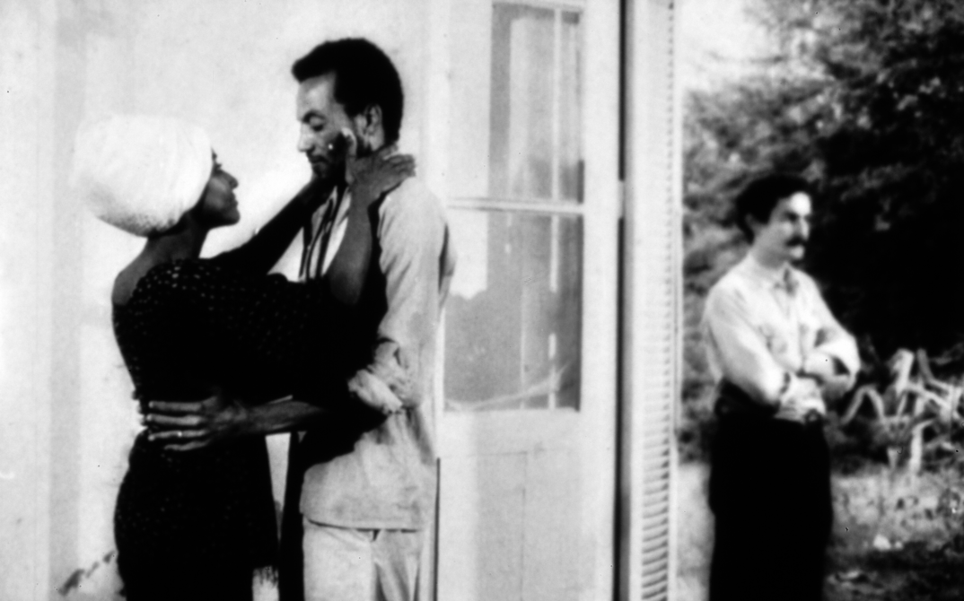 Filmstill aus MONANGAMBEE: Ein Mann und eine Frau in einer Umarmung. Im Hintegrund steht eine weitere Person.