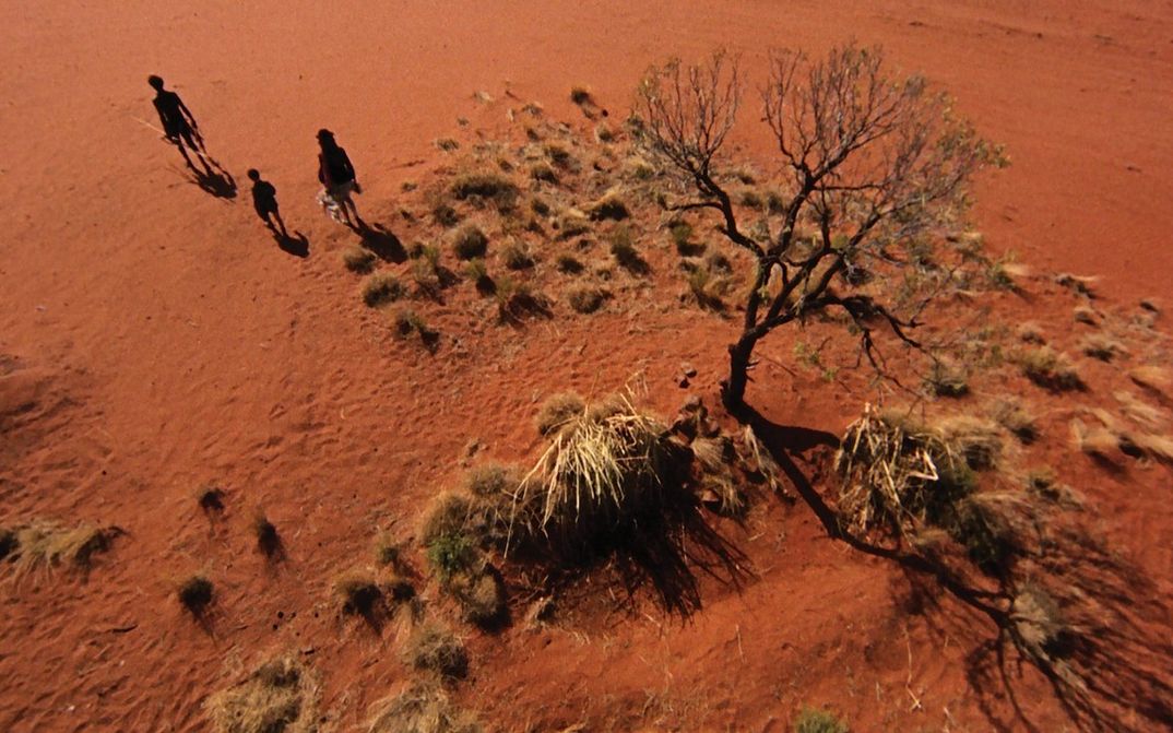 Film still from WALKABOUT: An aerial view of a desert with red sand. Three people can be seen at the edge of the picture.