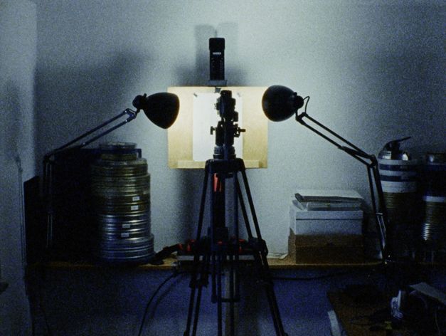 Filmstill aus "Spuren von Bewegung vor dem Eis" von René Frölke. Zu sehen ist ein dunkler Raum mit Filmrollen. In der Mitte steht ein Stativ, welches von zwei Lampen beleuchtet wird. 