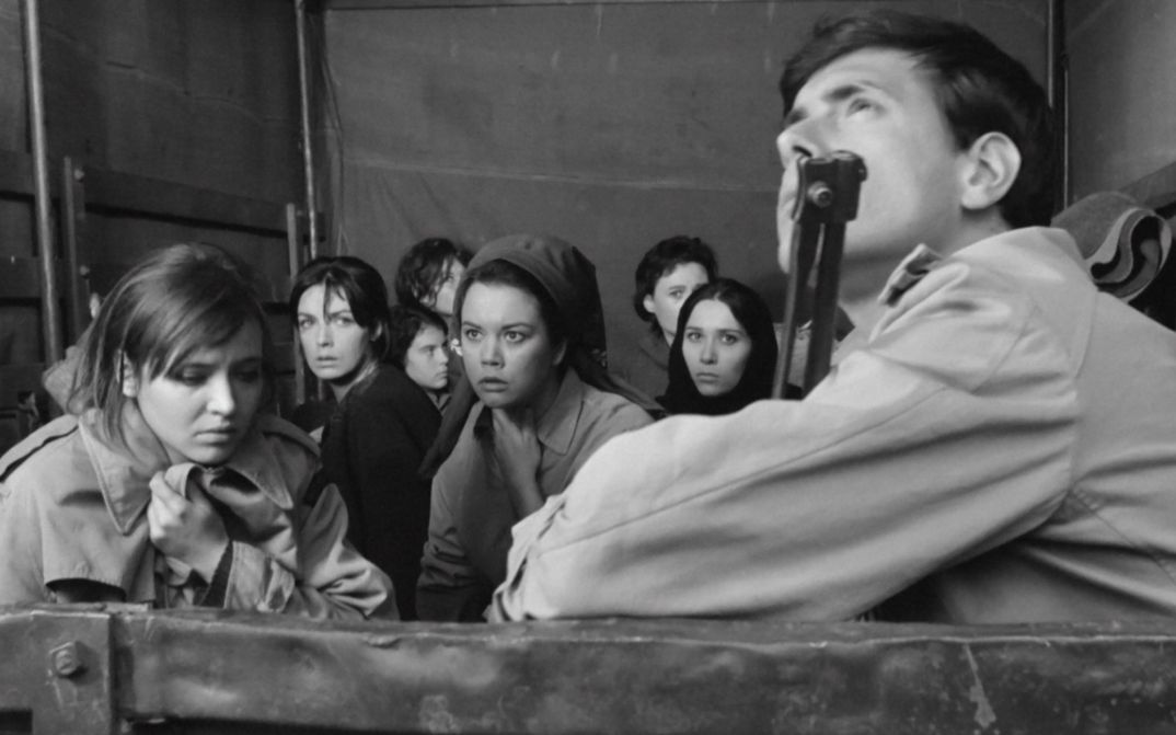 Filmstill aus LE SOLDATESSE: Mehrere Frauen mit verschrecktem Gesichtsausdruck sitzen auf einem Lastwagen. Im Vordergrund ist ein Soldat mit Gewehr.