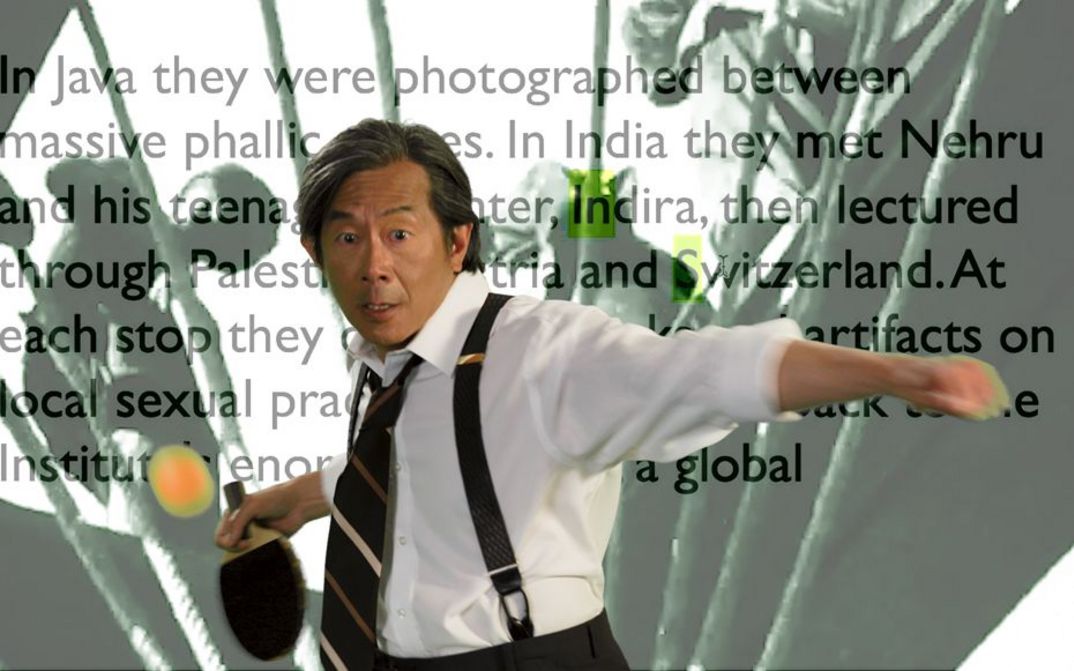 Film still from John Greyson’s film “Death Mask”. A person wears suspenders, a black striped tie and a white shirt, and is about to shoot a ball with a table tennis bat in front of a text about Java.