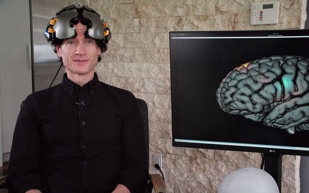 Film still from THEATRE OF THOUGHT: A man dressed in black with a helmet, next to him a monitor with the image of a brain.