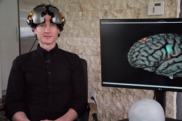 Film still from THEATRE OF THOUGHT: A man dressed in black with a helmet, next to him a monitor with the image of a brain. 