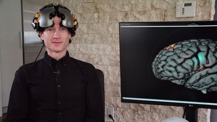 Film still from THEATRE OF THOUGHT: A man dressed in black with a helmet, next to him a monitor with the image of a brain. 