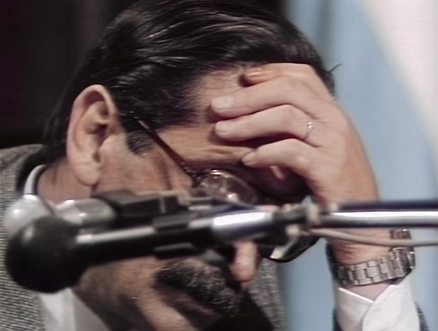 Still from the film "El juicio" by Ulises de la Orden. A blurry microphone can be seen in the foreground. Behind the microphone, a man with dark glasses and a dark moustache is resting his head on his hands.