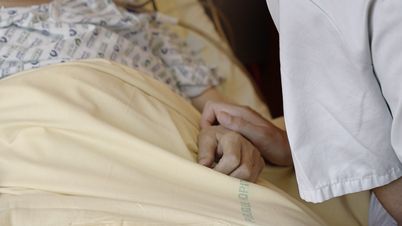 Still from the film „Notre corps“ by Claire Simon. A person lying in a bed. On the left is a person holding the hand of the lying person.