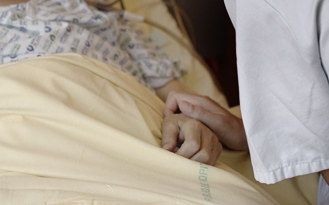 Still from the film „Notre corps“ by Claire Simon. A person lying in a bed. On the left is a person holding the hand of the lying person.