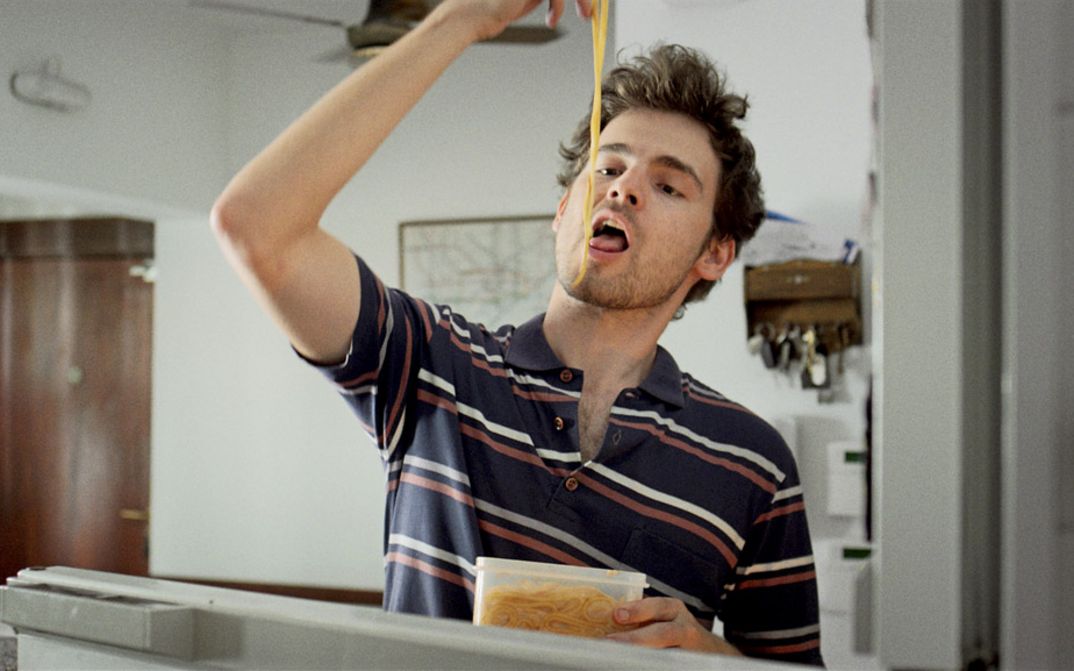 Filmstill aus „Arturo a los 30" von Martín Shanly. Ein junger Mann isst mit seinen Händen Nudeln aus einem Tupperdose.