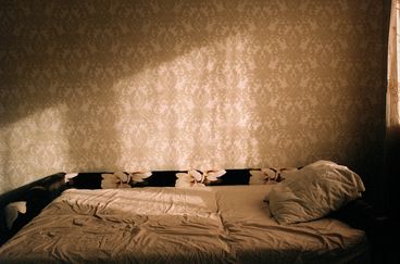 An empty futon against patterned wallpaper. There is a dynamic shadow from the window to the right of the frame, and the surface of the futon sheets are slightly crumpled.