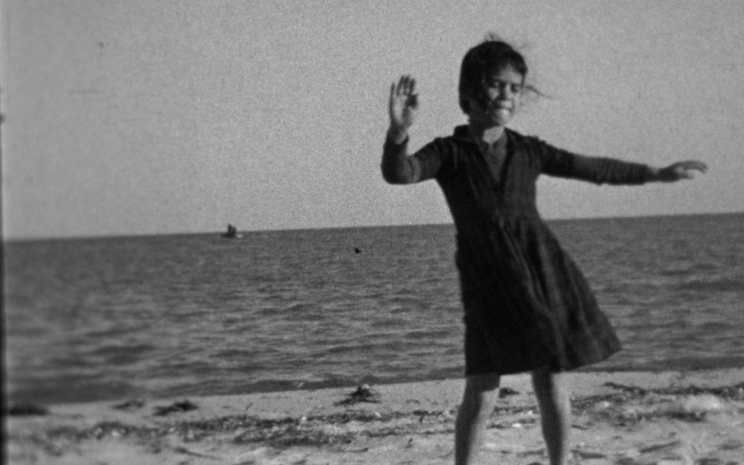 Filmstill aus BALLADE AUX SOURCES: Ein Mädchen in einem Kleid tanzend am Strand, hinter ihr das Meer.