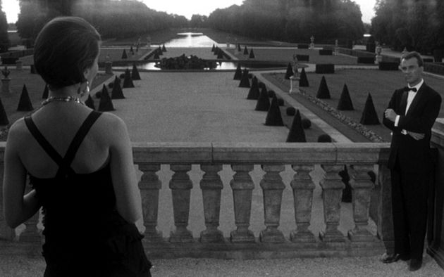 Still from the film "Last Year in Marienbad. A black and white image overlooking a terrace and a garden of a fancy villa. On the terrace, we see a woman on the left, she is wearing an evening gown, her back is turned away from the camera. On the right, a man in a tuxedo is looking at her.