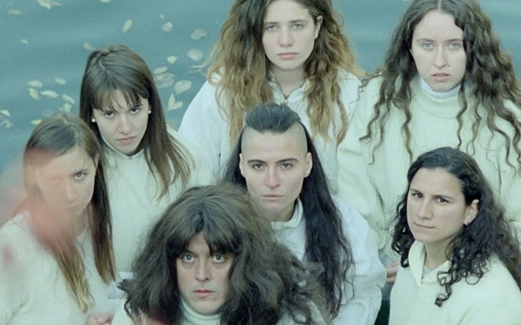 Filmstill aus „Mammalia“ von Sebastian Mihăilescu. Zu sehen ist eine Gruppe von sieben Personen mit langen Haaren aus der Vogelperspektive. Sie blicken alle in die Kamera. Im Hintergrund liegen Laubblätter.