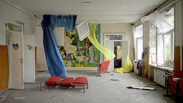 Filmstill from "Intercepted" by Oksana Karpovych. It shows an empty room with broken walls and dirt on the floor. Blue and yellow curtains hang in the room and there are red movie theater seats. 