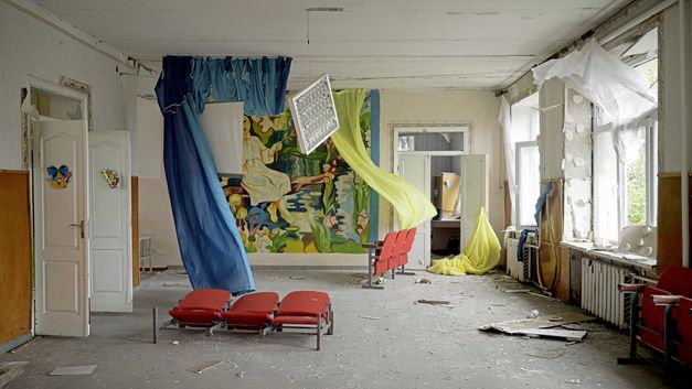 Filmstill aus "Intercepted" von Oksana Karpovych. Zu sehen ist ein leerstehender Raum mit kaputten Wänden und Schmutz auf dem Boden. In dem Raum hängen blaue und gelbe Vorhänge und es stehen rote Kinosessel.