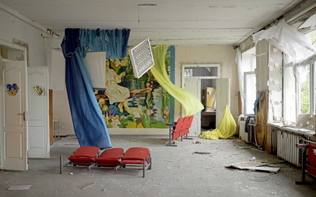 Filmstill aus "Intercepted" von Oksana Karpovych. Zu sehen ist ein leerstehender Raum mit kaputten Wänden und Schmutz auf dem Boden. In dem Raum hängen blaue und gelbe Vorhänge und es stehen rote Kinosessel.